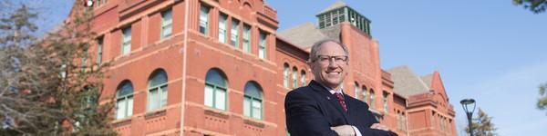 President Darrin 好 stand with arms crossed and smiling in front of the Old Main building.